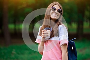 Portrait of beautiful brunette girl in sunglasses walking down the street. Keeping takeaway drink in hand. Smiling. Urban city