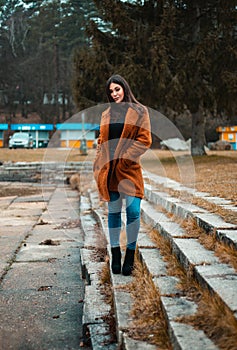 Portrait of beautiful brunette girl woman in orange fashion coat in season - photo on stairs with park and nature on background