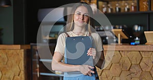 Portrait of the beautiful brunette waitress in apron with tablet in hands smiling cheerfully at camera in the cafe