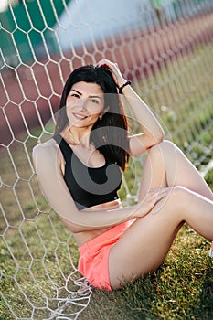 Portrait of Beautiful brunette woman in black top and pink shorts sitting on the lawn near a football goal at the stadium at