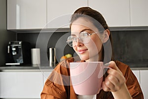 Portrait of beautiful brunette woman, sitting in kitchen, taking a break for cup of coffee. Girl drinks tea at home