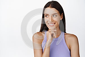 Portrait of beautiful brunette woman smiling, touching her chin thoughtful, looking aside at promo text, white