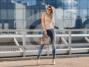 Portrait of beautiful brunette young woman in nice beige shirt, denim jeans and sunglasses. Genuine leather bag, high heels shoes