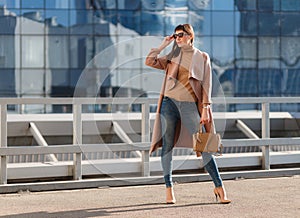 Portrait of beautiful brunette young woman in nice brown beige coat, denim jeans and sunglasses. Genuine leather bag, high heels