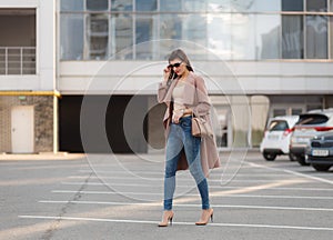 Portrait of beautiful brunette young woman in nice brown beige coat, denim jeans and sunglasses. Genuine leather bag, high heels