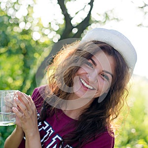 portrait of beautiful brunette young woman in white hipster hat happy smiling holding glass cup of water on green summe