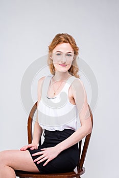 Young beautiful woman with long red, curly hair sitting on a wooden chair on a white background in the studio. Dressed in a white