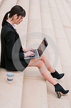 Portrait of beautiful businesswoman using laptop sitting on stairs in city street.