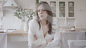 Portrait of beautiful caucasian girl sitting in stylish kitchen and thinking. Pensive brunette young woman in white