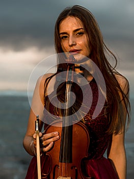 Portrait of beautiful Caucasian woman holding violin on the beach. Music and art concept. Girl wearing red dress in nature. Sunset