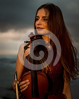 Portrait of beautiful Caucasian woman holding violin on the beach. Music and art concept. Girl wearing red dress in nature. Sunset