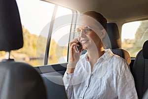 Portrait of beautiful charming cute businesswoman wearing glasses and white shit sitiing in taxi and talking on cell phone and