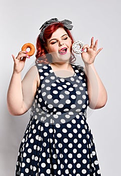 Portrait of beautiful cheerful fat plus size woman pin-up wearing a polka-dot dress isolated over light background, eating a donut