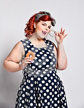 Portrait of beautiful cheerful fat plus size woman pin-up wearing a polka-dot dress isolated over light background, eating a donut