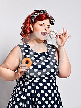 Portrait of beautiful cheerful fat plus size woman pin-up wearing a polka-dot dress isolated over light background, eating a donut