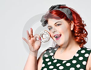 Portrait of beautiful cheerful fat plus size woman pin-up wearing a polka-dot dress isolated over light background, eating a donut