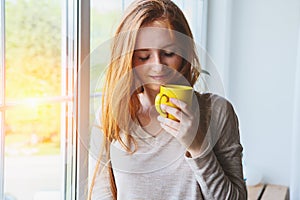 Portrait of beautiful cheerful young smiling red hair woman holding yellow coffee cup, enjoying hot morning coffee at sunrise morn
