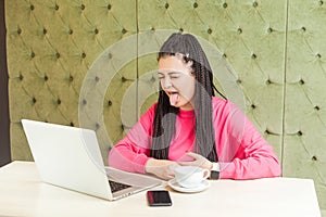 Portrait of beautiful childish funny young girl freelancer with black dreadlocks hairstyle in pink blouse sitting and showing