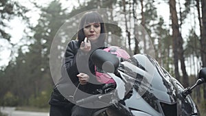 Portrait of beautiful confident motorbiker applying red lipstick outdoors looking in side mirror. Brunette Caucasian