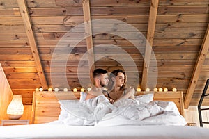 Portrait of beautiful couple embracing and sitting on bed under white blanket. Relax at home in cozy wooden bedroom