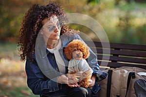 Portrait of beautiful curly haired woman holds small dog on her hands, while sitting in autumn park. Outdoor pet. Toy