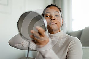 Portrait of beautiful curvy woman, looking at small round makeup mirror at home.