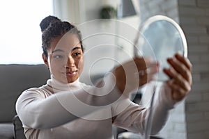 Portrait of beautiful curvy woman, looking at small round makeup mirror at home.