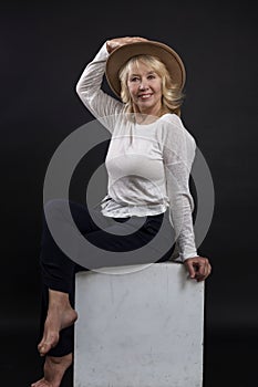 Portrait of a beautiful elderly woman. A smiling blonde girl in a hat gracefully sits on a white cube. Happy maturity. Close-up.