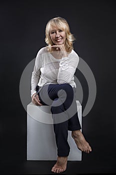 Portrait of a beautiful elderly woman. A smiling blonde sits gracefully on a white cube. Happy maturity. Close-up. Black