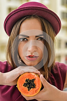 Portrait of beautiful elegant female wearing red felt hat holding half of papaya fruit next to her face