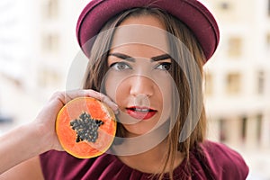 Portrait of beautiful elegant female wearing red felt hat holding half of papaya fruit next to her face