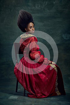 Portrait of beautiful, elegant young african woman, medieval princess in vintage dress sitting, posing against dark
