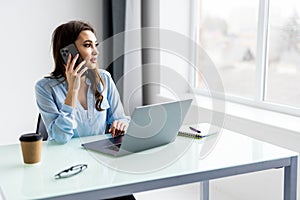 Portrait of beautiful executive woman making call while sitting at her workplace in front of laptop and working on new project