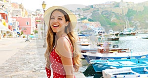 Portrait of beautiful fashion girl in Procida, Italy. Attractive young woman with red dress and hat walks along the harbor of