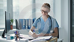 portrait of beautiful female medical doctor wearing blue uniform with stethoscope, smiling woman in glasses writes in