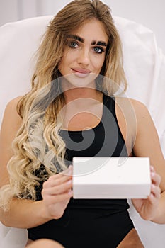 Portrait of beautiful female model in cosmetology studio hold white box in a hand. Sexy woman in black bodysuit