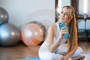 Portrait of beautiful fitness woman sitting on mat with sports bottle in her hands.
