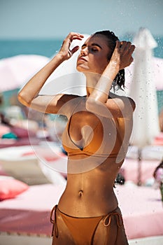 Portrait of beautiful girl in beige bikini dreamily closing her eyes while taking shower on beach. Young pretty girl in