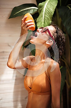 Portrait of beautiful girl in beige bikini and sunglasses standing and squeezing grapefruit juice with hand