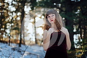 Portrait of a beautiful girl in a dark long dress who stands alone among the trees in the winter forest