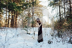 Portrait of a beautiful girl in a dark long dress who stands alone among the trees in the winter forest