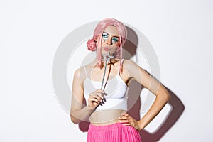 Portrait of beautiful girl in fairy costume looking thoughtful, holding magic wand and wearing pink wig, standing over