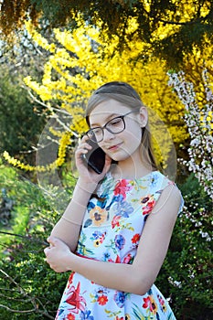 Portrait of beautiful girl with glasses and brown hair talk on smart phone. Young smiling teneeger in green spring park resting