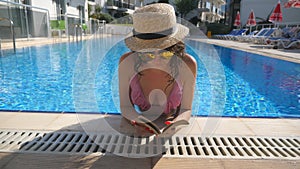 Portrait of beautiful girl in hat and sunglasses reading book at pool. Young woman relaxing at warm sunny day during