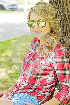 Portrait of a beautiful girl with large plump lips curls in denim shorts and a shirt in sunglasses sitting near a tree