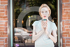 Portrait beautiful girl with long blonde hair holds a coffee cup stands by the window of a street city cafe, looks to camera