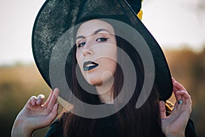 Portrait of a beautiful girl with pale skin and black lips in a big black hat. Woman in the image of a witch for Halloween.