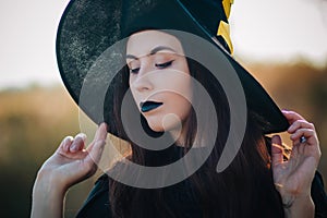 Portrait of a beautiful girl with pale skin and black lips in a big black hat. Woman in the image of a witch for Halloween.