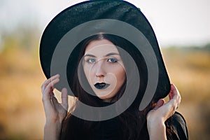 Portrait of a beautiful girl with pale skin and black lips in a big black hat. Woman in the image of a witch for Halloween.