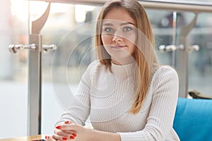 Portrait of beautiful girl with perfect skin. Blonde female looks at camera with crossed fingers, spends her free time in cafe,